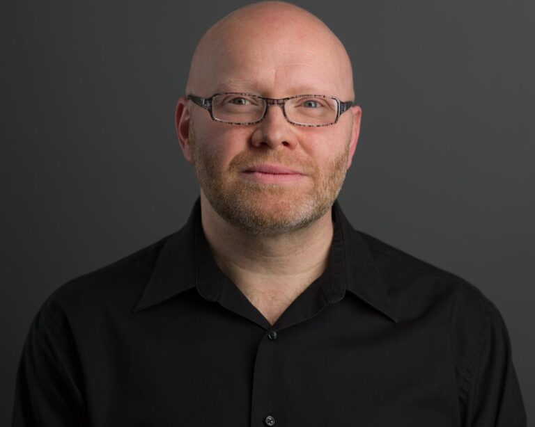 A bald man with a light beard wearing glasses and a black collared shirt poses against a neutral gray background.