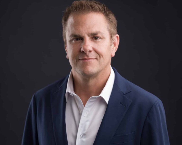 A professional portrait of a man wearing a navy blue suit and white shirt, standing against a dark background. He has short hair and is smiling slightly, conveying confidence and approachability.