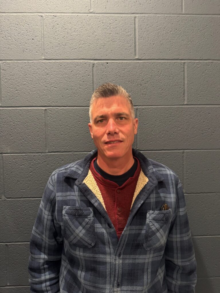 A man with short hair is standing against a grey brick wall. He is wearing a plaid jacket with a red shirt underneath, and there appears to be a pen in his left chest pocket.