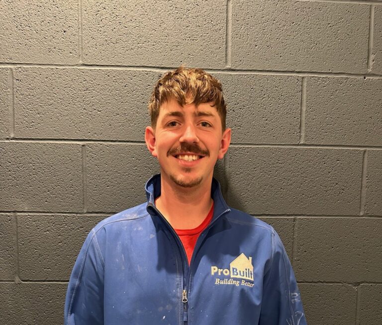 A man with short brown hair and a mustache, smiling while wearing a blue jacket with a logo that says 'Pro Built Building Better'. He is standing against a textured grey brick wall.