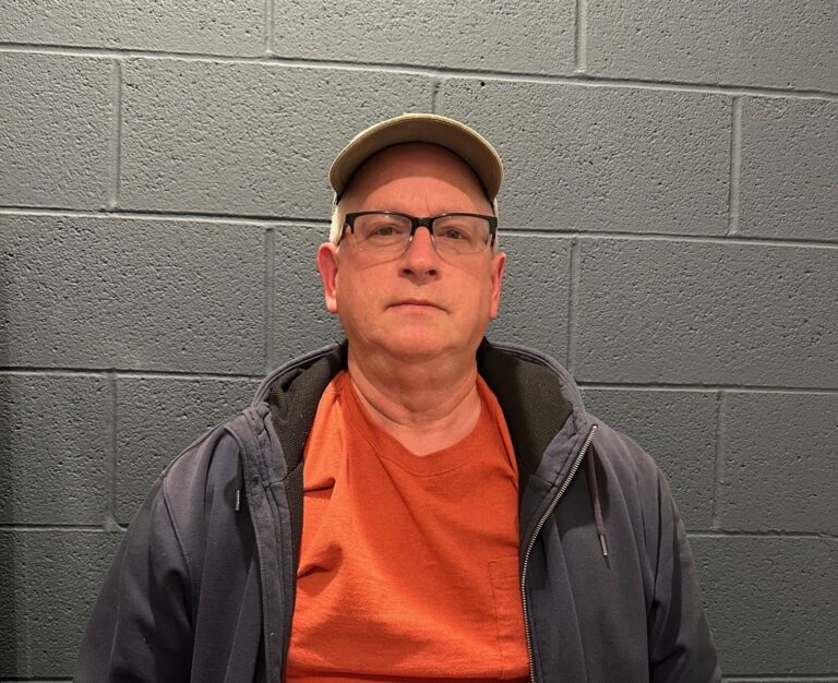 A middle-aged man wearing glasses, a beige cap, an orange shirt, and a dark jacket, standing in front of a gray brick wall.