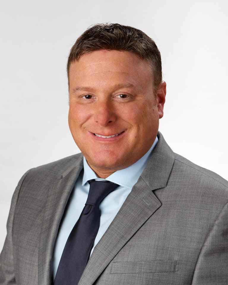 A professional portrait of a man wearing a gray suit, blue shirt, and dark tie. He is smiling and has short brown hair, set against a plain gray background.