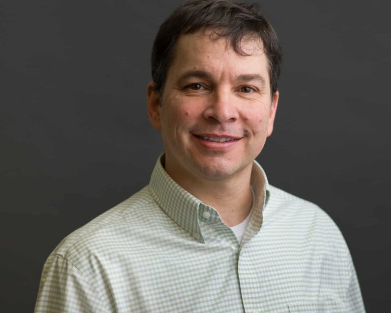 A man with dark hair wearing a light green checkered shirt smiles while standing against a plain gray background.