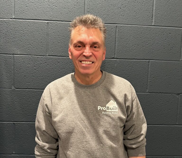 A man smiling, wearing a gray sweatshirt with a logo that reads 'ProBuilt Building Better', stands in front of a textured gray brick wall.