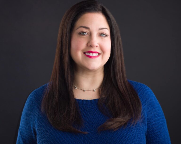 A woman with long dark hair and a bright smile is wearing a blue textured sweater. She stands against a dark background, looking directly at the camera. Her makeup is subtle, and she is wearing a small necklace.
